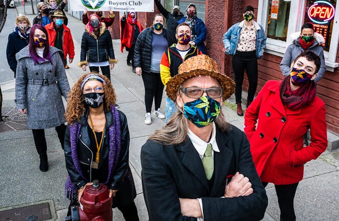 A group of people wearing coloured face masks stands on a sidewalk facing the cam.