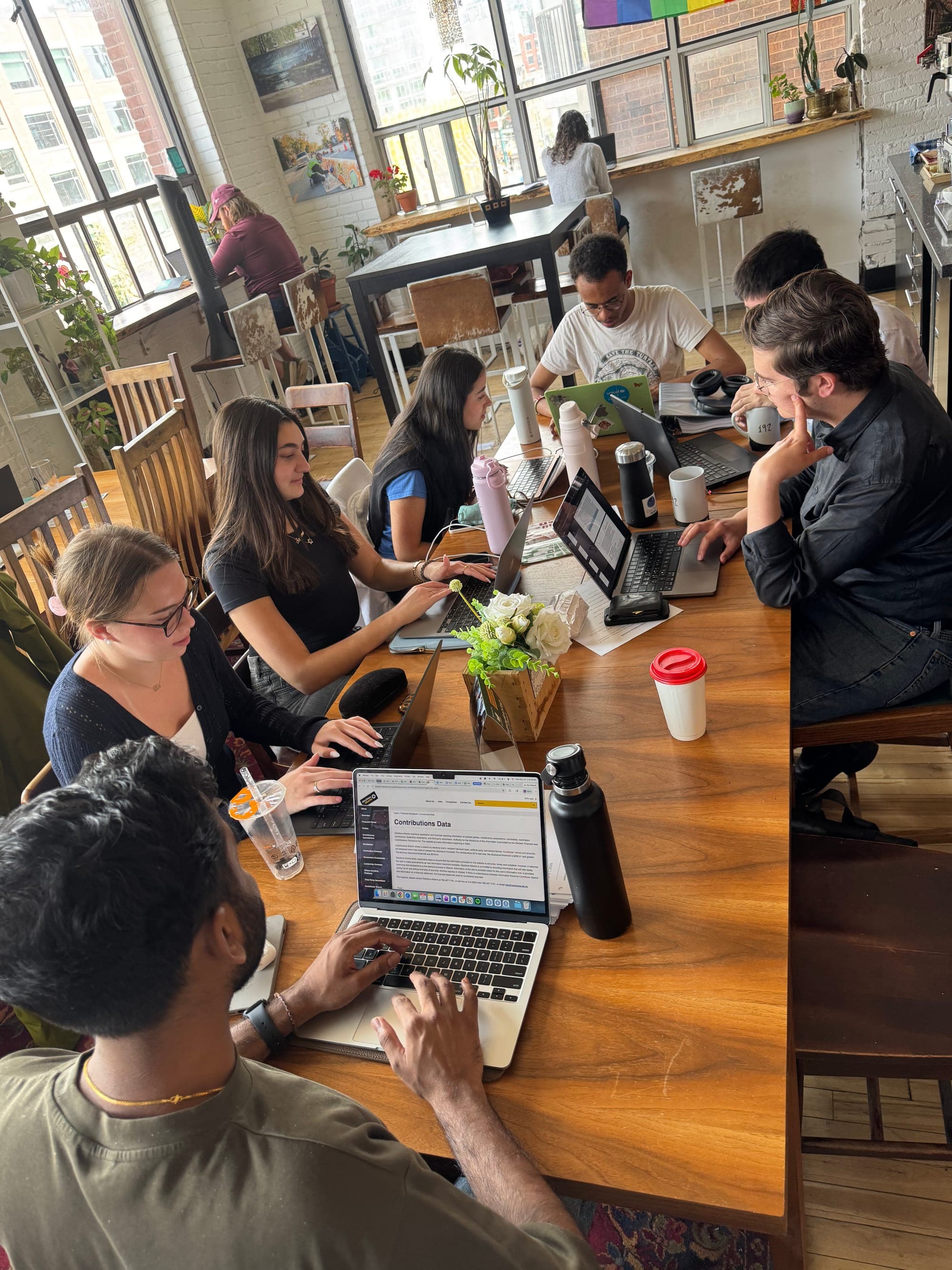 A group of seven students all turn towards their computers while working on projects for their internship at a long wooden table in an office setting.