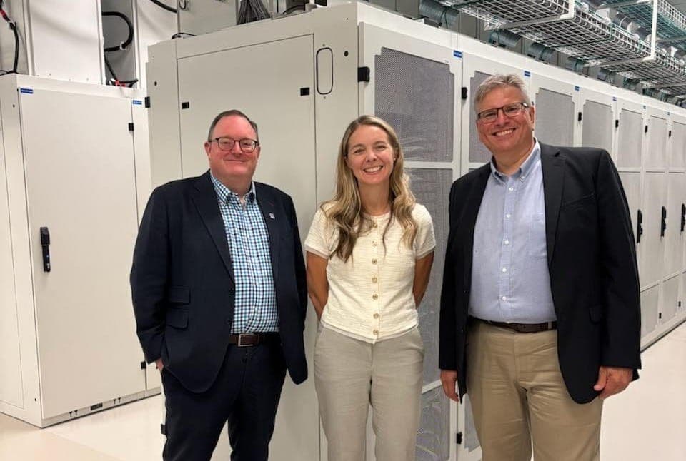 Three people stand in front of components of a data centre.