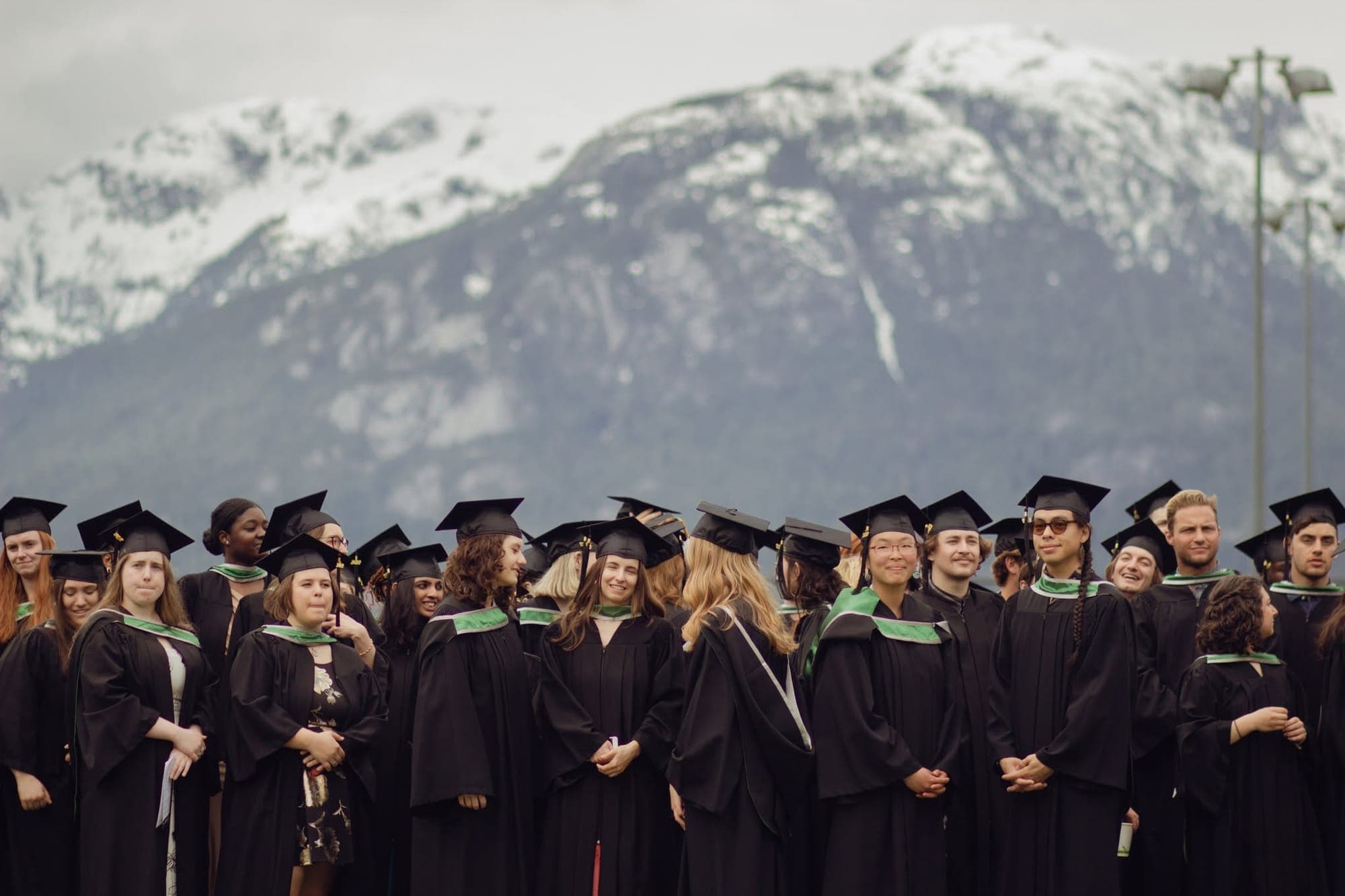 A group of students wearing graduations caps and gowns smile at the camera and chat with one another in front of a snowy mountain.