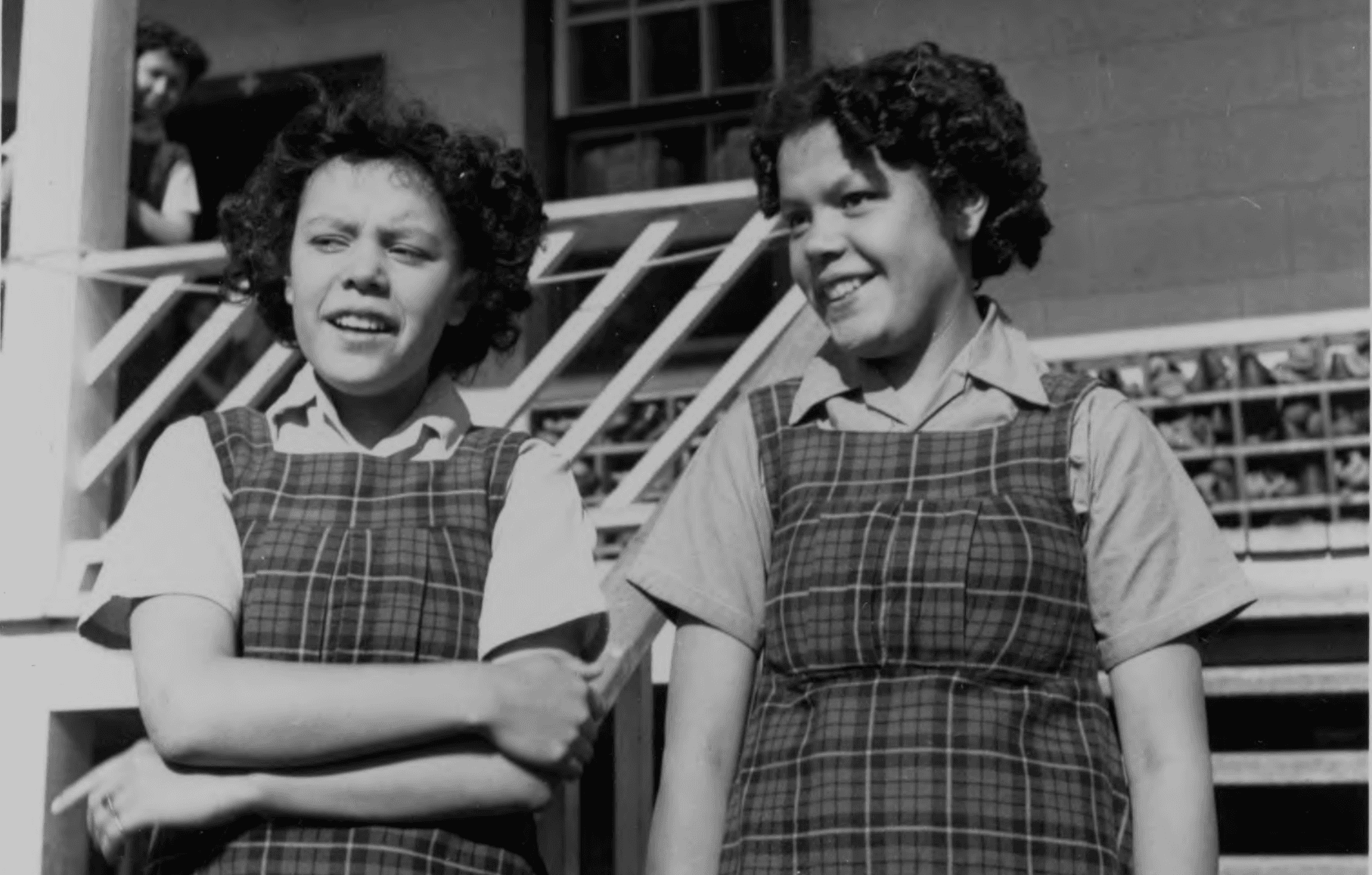 Two Indigenous girls with short curled hair and plaid school uniforms stand in front of Sturgeon Lake Residential School in Alberta.