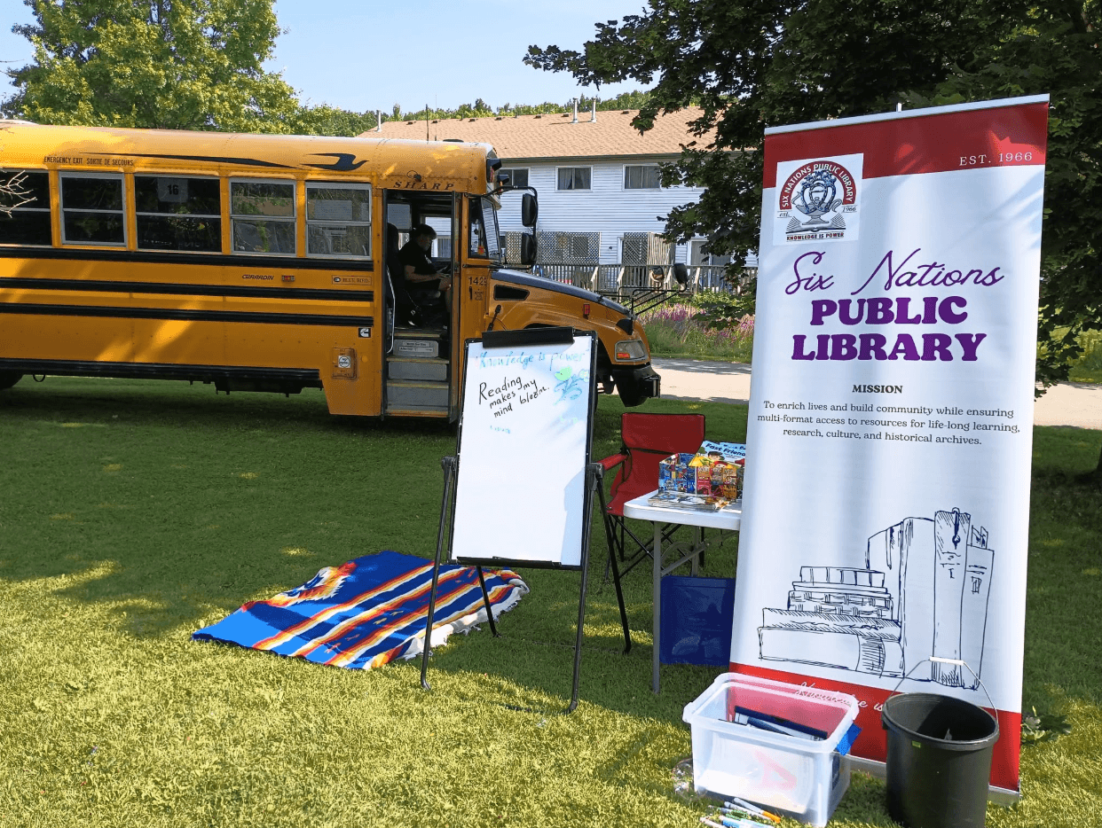 A sign for the Six Nations Public Library is seen on a lawn in front of a school bus.