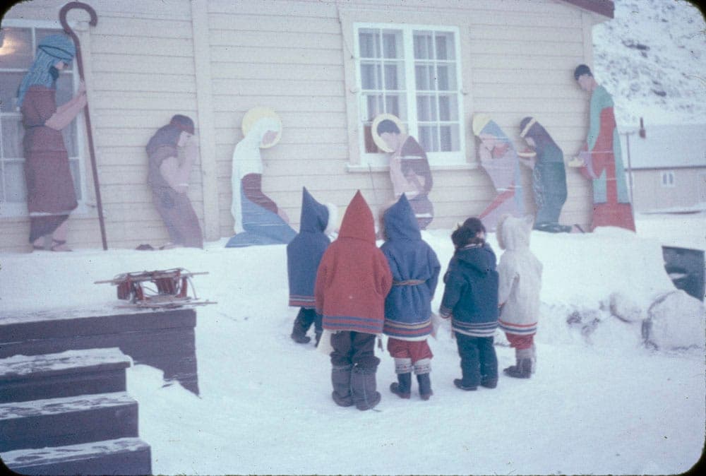 Five children stand outside the Pangnirtung Federal Day School, facing a wooden nativity scene laid across the front of the building.
