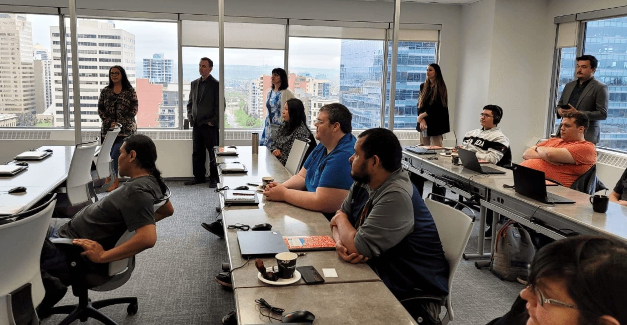 A group of people sit at tables in front of laptops in an office building.