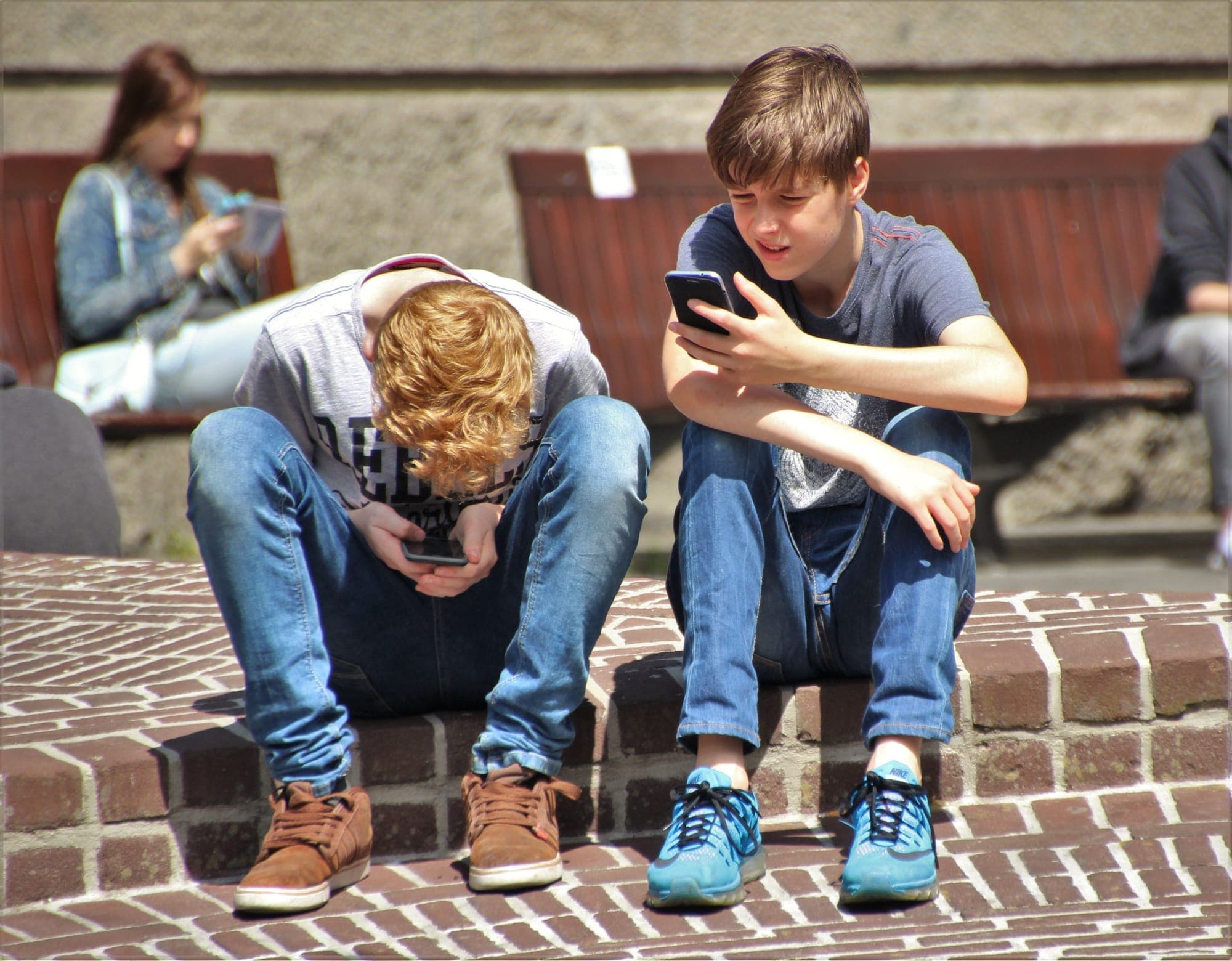 Two teenage boys are sitting in a public area looking at their phones
